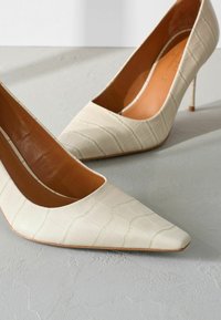 Décolleté in pelle verniciata beige con texture coccodrillo, punta appuntita e sottile tacco a stiletto alto. L'interno presenta una fodera liscia color camel.