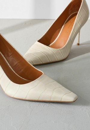 Bēšas laku pēdas ar krokodila tekstūru, ar smailu purngalu un plānu, augstu stiletto papēdi. Iekšpusē ir gluda brūna odere.