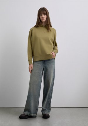 Hanorac verde măsliniu cu buzunar tip kangur, asortat cu blugi largi de culoare albastru deschis și pantofi negri masivi cu platformă. Design minimalist, textură moale.