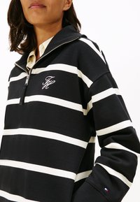 Melnbalts svītrains zip-up džemperis ar augstu apkakli, dekorēts ar izšūtu logotipu un mazu karodziņa detaļu uz piedurknes.