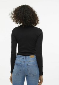 Eine Frau mit lockigem Haar, die ein schwarzes Langarmoberteil und blaue hochtaillierte Jeans trägt, steht mit dem Rücken zu einem weißen Hintergrund.