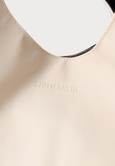 Beżowa torba tote z gumy o gładkiej fakturze i zaokrąglonym otworem. Posiada wytłoczone logo "STUTTERHEIM" oraz kontrastowe szwy.