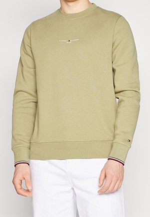 Beige Tommy Hilfiger-sweatshirt met klein logo op de borst en mouw, gedragen door persoon in witte broek tegen een effen achtergrond.
