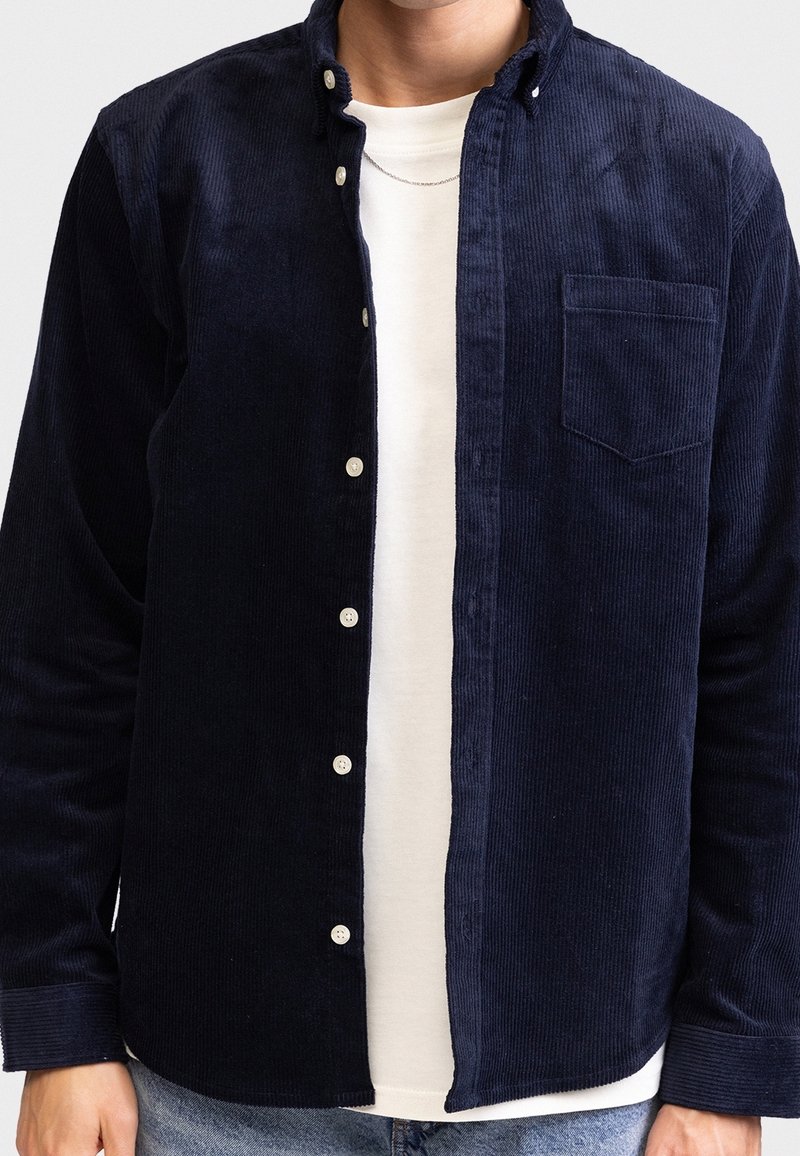 Man in een onknopte donkerblauwe ribfluwelen shirt over een witte crewneck t-shirt en lichtblauwe jeans, halverwege de nek en taille geknipt.