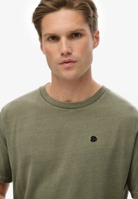 Oliwkowa bawełniana koszulka z okrągłym dekoltem i krótkimi rękawami. Zawiera małe czarne logo haftowane na klatce piersiowej. Miękka tekstura.