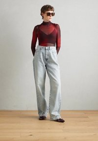 Haut transparent rouge à manches longues avec un design superposé, jeans taille haute bleu clair, et mocassins marron foncé, debout sur un sol en bois.