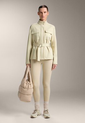 Nő, aki krém színű, öves dzsekit, bézs leggingseket, fehér zoknit és tornacipőt visel, és egy bézs pufi táskát tart egyszerű háttér előtt.