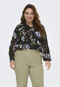 Blusa nera a fiori con fiori blu e verdi, maniche lunghe, scollatura a V, abbinata a pantaloni cargo beige chiaro con tasche laterali.