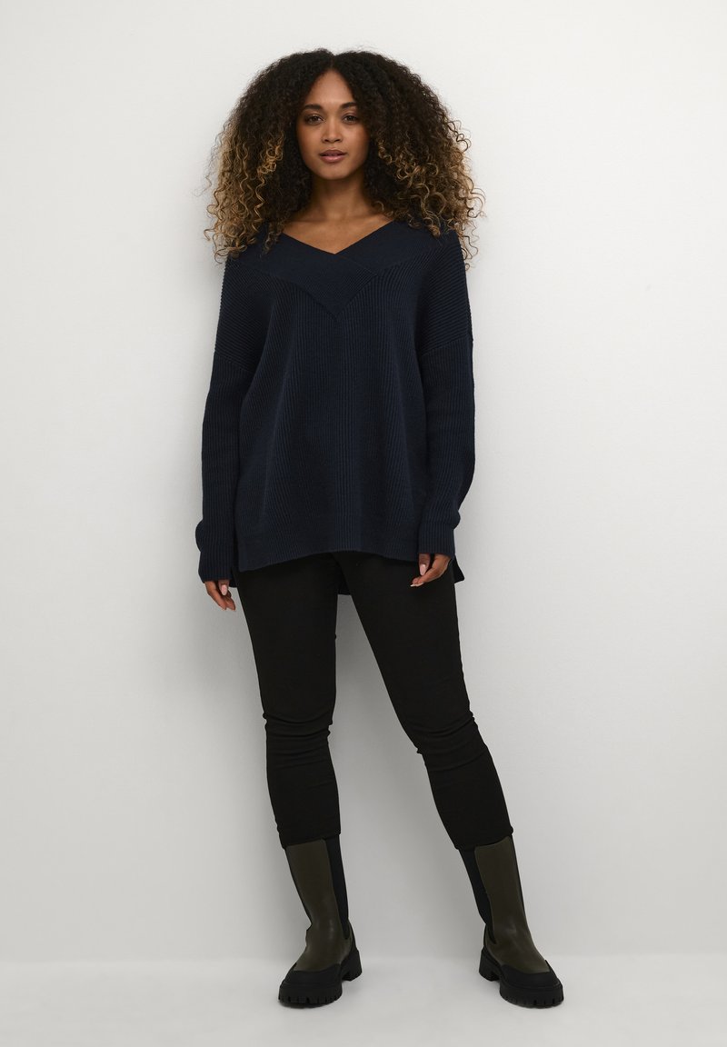 Pulover oversize de tricot cu decolteu în V de culoare bleumarin închis, cu textură în dungi, asortat cu pantaloni negri estruturati și cizme negre de gleznă verde închis.