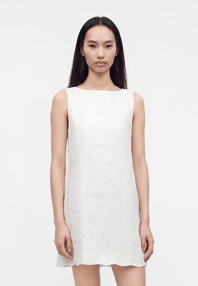 Woman with long straight black hair wearing a sleeveless white linen dress standing against a plain light background.