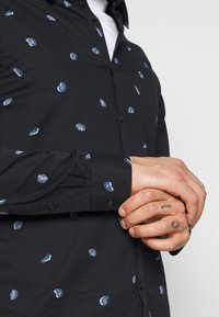 Mani unite davanti a una camicia blu navy con piccoli motivi di conchiglie bianche e blu, con un tatuaggio visibile sul dito medio destro.