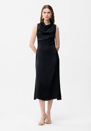 Kobieta w czarnej, bezrękawnikowej sukience midi z marszczonym dekoltem i złotych sandałach na obcasie, stoi z rękami za plecami.
