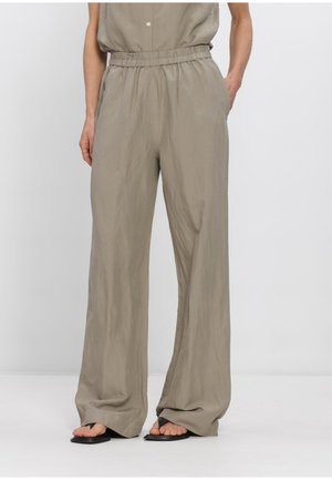 Vrouw die loszittende, hooggetailleerde taupekleurige broek draagt met elastische tailleband en zijzakken, gecombineerd met zwarte teenslippers.