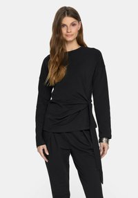 Czarny top z długim rękawem, z charakterystycznym skręconym przodem. Gładki materiał, okrągły dekolt, w zestawieniu z czarnymi spodniami o luźnym kroju. Prosty design.