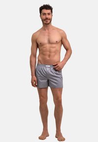 Boxer rayé gris avec une ceinture élastique. Le tissu semble lisse avec une coupe décontractée, le modèle pose pieds nus dans un cadre neutre.