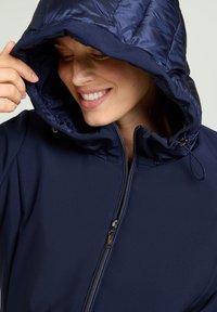 Marineblauwe gewatteerde jas met glanzend materiaal op de capuchon en een gladde stoffen body. Voorzien van een ritssluiting en verstelbare capuchonkoorden.
