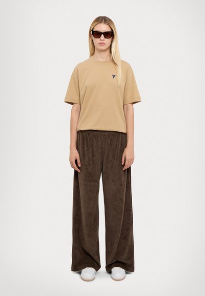 Beige oversized t-shirt met een klein blauw logo, gecombineerd met bruine wijde broek gemaakt van zachte stof. Witte sneakers maken de outfit compleet.