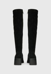 Botas altas de ante negro hasta la rodilla con tacones de bloque, vistas desde la parte de atrás, que presentan cremalleras laterales y una textura suave.