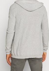 Šedý pletený kardigan s kapucí, vyrobený z texturované látky, s žebrovanými manžetami a bočními rozparky pro pohodlný střih.