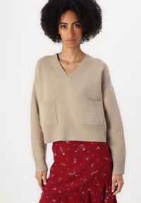 Beige pletený svetr s výstřihem do V, který má dvě přední kapsy a decentní logo. Kombinovaný s červenou sukní s květinovým vzorem.