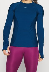Długi rękaw sportowej bluzki w ciemnym niebieskim kolorze, z teksturowanymi bocznymi panelami i okrągłym dekoltem z małym logo na piersi. W połączeniu z fioletowymi legginsami.