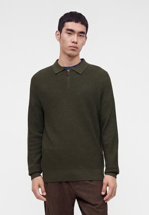Giovane uomo con maglione verde scuro texturizzato con zip al collo e pantaloni marroni a quadri, in piedi su sfondo bianco uniforme.