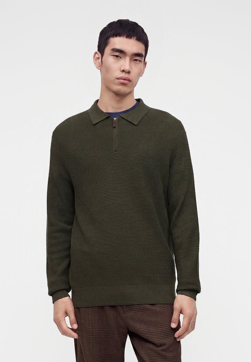 Jeune homme portant un pull avec col zippé texturé vert foncé et un pantalon marron à carreaux, debout sur un fond blanc uni.