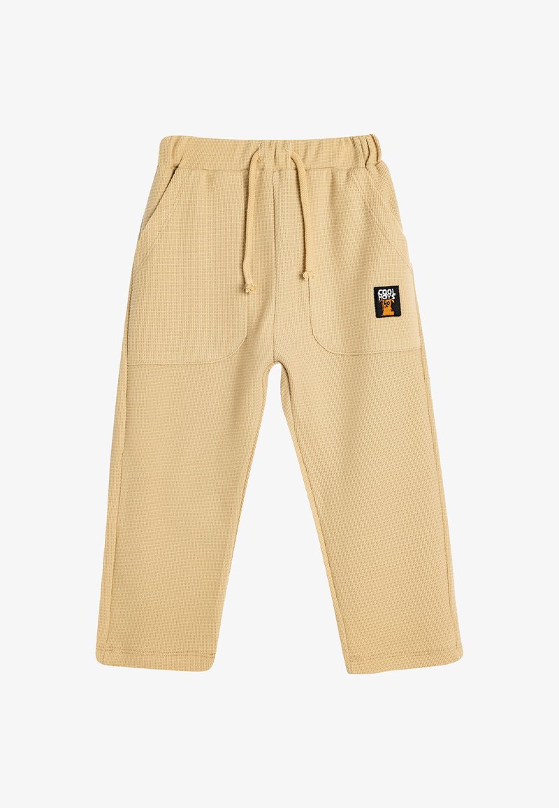 Beige bukser med vaffelstruktur, elastisk talje og snor, der har to sidelommer og et lille logo-patch foran.