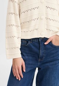Krämfärgad stickad cardigan med horisontella utskärningsmönster, sex knappar och långa ärmar, tillsammans med mörkblå denimjeans.