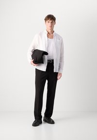 Camisa de linho branca sobre uma camisola de alças branca, calças pretas, segurando uma peça de vestuário preta enrolada. Sapatos pretos com sola grossa.
