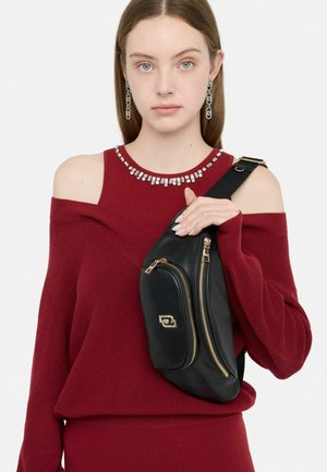 Schwarze Leder-Umhängetasche mit goldenen Reißverschlüssen und einem geschwungenen Design. Modell trägt einen tiefroten, schulterfreien Pullover mit einem Perlenkragen.