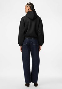 Felpa nera con cappuccio dal fit morbido, abbinata a jeans a gamba larga blu scuro, che mostrano uno stile casual e contemporaneo.