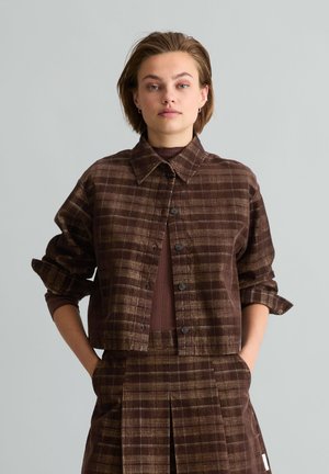 Junge Frau mit kurzen Haaren, die eine braune karierte Jacke und einen passenden Rock trägt, die Hände in den Taschen, vor einem schlichten hellen Hintergrund.