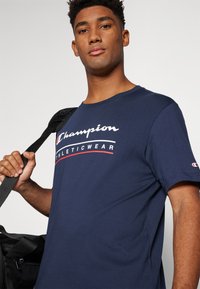 Navy blue cotton t-shirt featuring "Champion Athleticwear" logo in white and red, crew neck, short sleeves, and a relaxed fit.