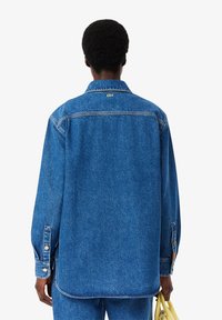 Blue denim shirt with a collar, long sleeves, and buttoned cuffs. Features a subtle logo on the back, with a slightly curved hem.