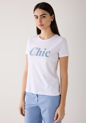Junge Frau mit langen dunklen Haaren, die ein weißes "Chic"-T-Shirt und hellblaue Hose trägt, steht vor einem schlichten hellen Hintergrund.