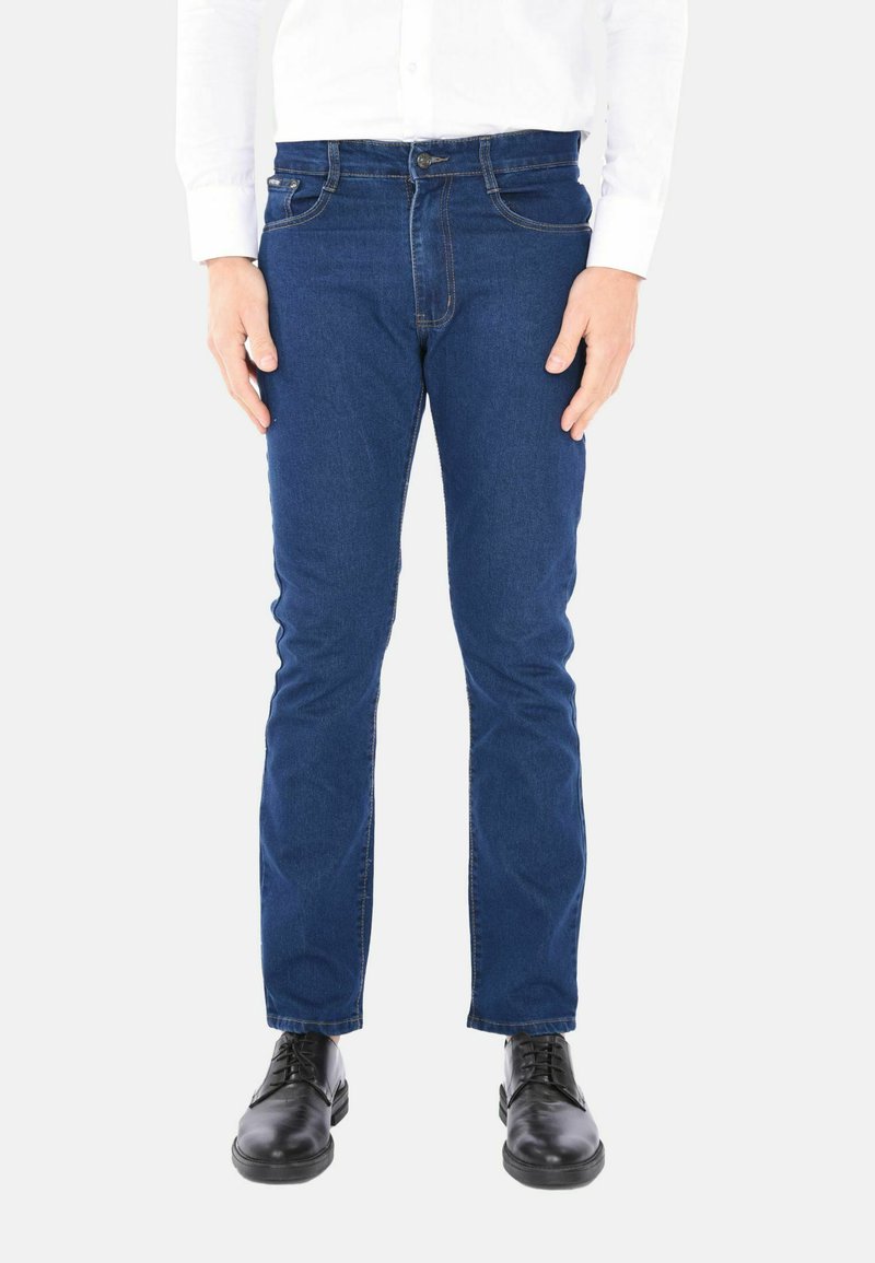 Uomo che indossa jeans di denim blu, una camicia bianca a maniche lunghe e scarpe eleganti in pelle nera, in piedi davanti a uno sfondo bianco.