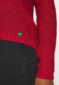 Roter gestrickter Pullover mit loser, texturiertem Gewebe; verfügt über gerippte Bündchen und Saum, kleiner grüner Anhänger mit der Aufschrift „ONLY“ nahe dem Saum.