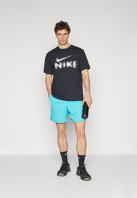 Camiseta negra de Nike con logo blanco, pantalones cortos azules, zapatillas negras y sosteniendo una botella de agua negra; tejido suave, corte relajado.