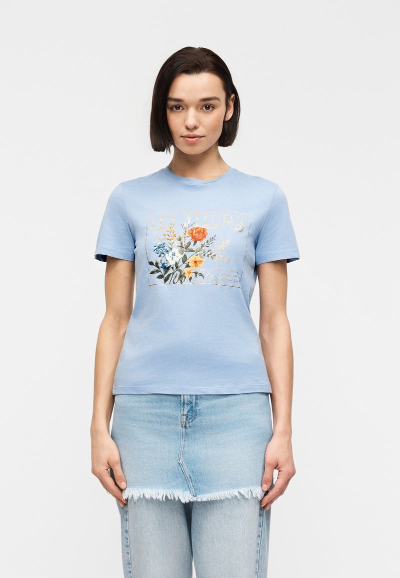 Femme portant un T-shirt bleu clair avec un imprimé floral coloré et le texte « Les Fleurs », associé à une jupe en jean clair superposée sur un jean.