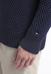 Gros plan sur une main reposant sur le côté, portant un pull en tricot texturé bleu marine et un pantalon beige.