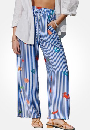 Damskie szerokie spodnie w niebiesko-białe paski z nadrukami o tematyce morskiej i talią ze sznurkiem, zestawione z białą koszulą i beżowymi sandałami.