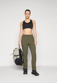 Pantalones de exterior en verde oliva con un ajuste slim, que cuentan con bolsillos con cremallera, combinados con un sujetador deportivo negro y una mochila negra y gris.