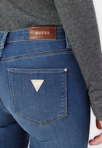Jeans din denim albastru cu un eticheta din piele maro pe talie, având o patch triangular pe buzunarul din spate drept.
