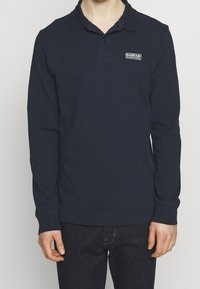 Osoba ubrana w granatową koszulę polo z długim rękawem z logo Barbour International na lewej piersi, w połączeniu z ciemnymi dżinsami.