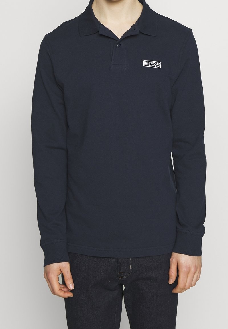 Osoba ubrana w granatową koszulę polo z długim rękawem z logo Barbour International na lewej piersi, w połączeniu z ciemnymi dżinsami.