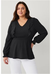 Haut noir à manches longues avec un col en V et des accents volantés, présentant une taille cintrée et un tissu texturé. Associé à un pantalon blanc.