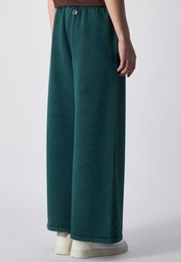Teal wijde broek gemaakt van zachte stof, met een elastische tailleband met een logo aan de voorkant en een relaxte pasvorm. Gestyled met witte sneakers.