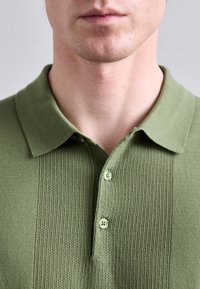 Hombre vestido con una camiseta polo de punto texturizado en verde oliva con dos botones abrochados, mostrado desde la barbilla hasta la parte superior del pecho sobre un fondo liso.