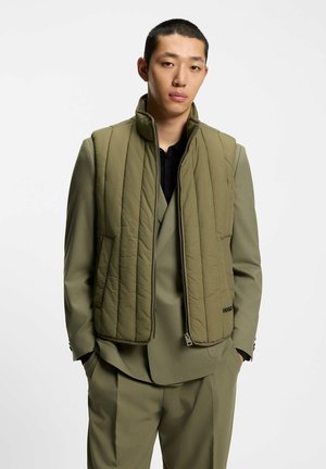 Giovane uomo con indosso un completo verde oliva abbinato a un gilet trapuntato, in piedi con le mani in tasca davanti a uno sfondo bianco uniforme.